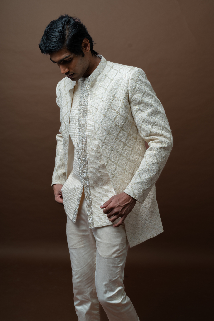 South Indian groom wearing designer ivory Jodhpuri suit with intricate detailing on brown backdrop.