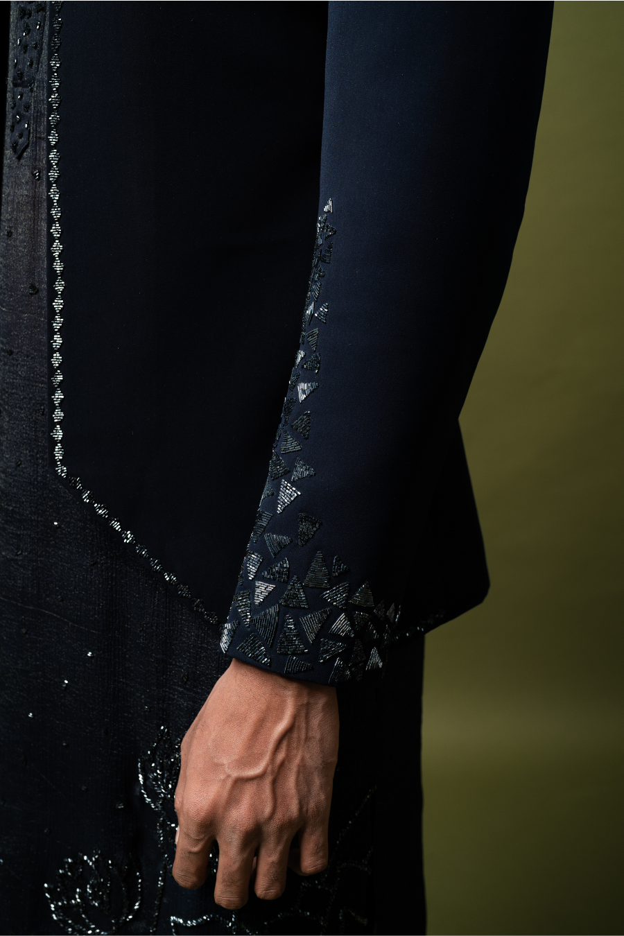 Man in a navy blue designer Jodhpuri suit, seated against olive backdrop, perfect for Indian wedding functions.
