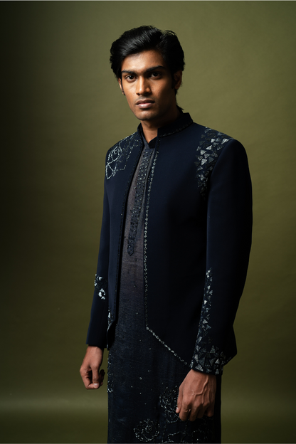 Full-sleeve navy blue Jodhpuri jacket paired with embroidered kurta, worn by a groom on a stool.

