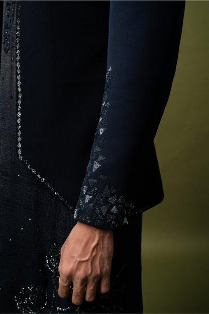 Man in a navy blue designer Jodhpuri suit, seated against olive backdrop, perfect for Indian wedding functions.


